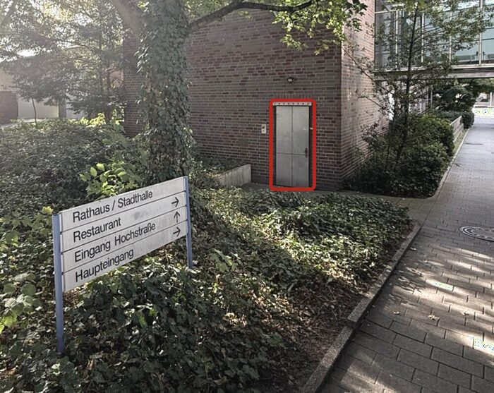 Blick auf das Backsteingebäude neben der Stadthalle Kamen. Die metallene Doppeltür, die zur Kegelbahn führt, ist rot umrandet. Links davor steht ein Wegweiser mit den Richtungen Rathaus und Stadthalle, Restaurant, Eingang Hochstraße und Haupteingang; rechts führt ein gepflasterter Weg am Gebäude entlang.